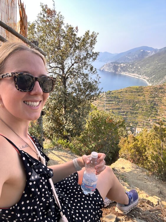 Klaudia sits on the ground and enjoys the sights from her trip between villages. She sees the sea and the green hills of Manarola.