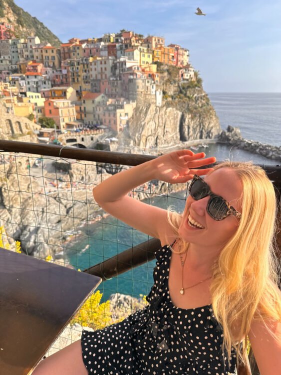 Klaudia, adorned in a polka-dotted black dress, sits in a restaurant chair, happily giggling. With a turquoise sea and yellow flowers in the foreground, Manarola village stands proudly in the background.
