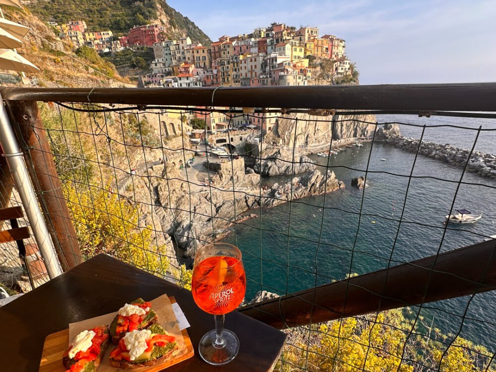 an amazing view of Manarola village in Cinque Terre, from a balcony of a restaurant, on the table is Aperol Spritz and bruschetta, below a sea and Manarola full of colorful little houses on the rocks