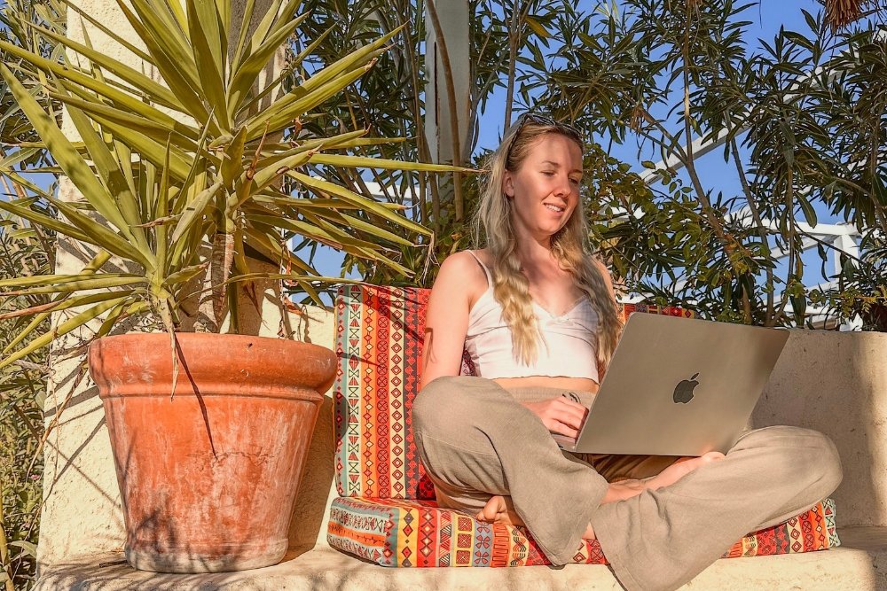 Klaudia sits cross-legged with her laptop on her lap in long linen pants and a white top. She sits outside in a restaurant with flowers and bushes behind her