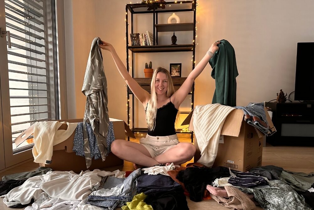 Between two boxes of clothes, Klaudia is sitting on the floor with her legs crossed. She is holding her used clothes in both hands raised up. Other clothes are lying loosely on the floor next to her.